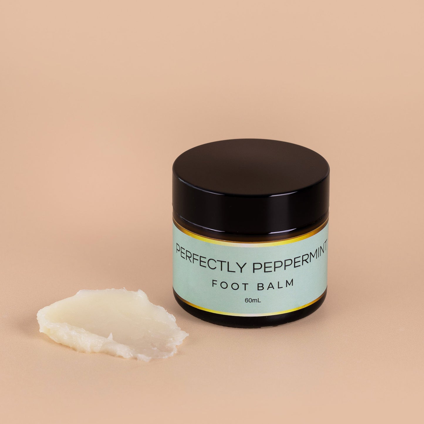 Nourishing Mana Perfectly Peppermint Foot Balm in a 60ml amber glass jar with a sample of the balm texture beside it on a beige background.