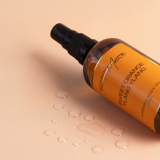 Nourishing Mana Sweet Orange Ylang Ylang Morning Body Oil in a 100ml amber glass spray bottle with oil droplets on a beige background.