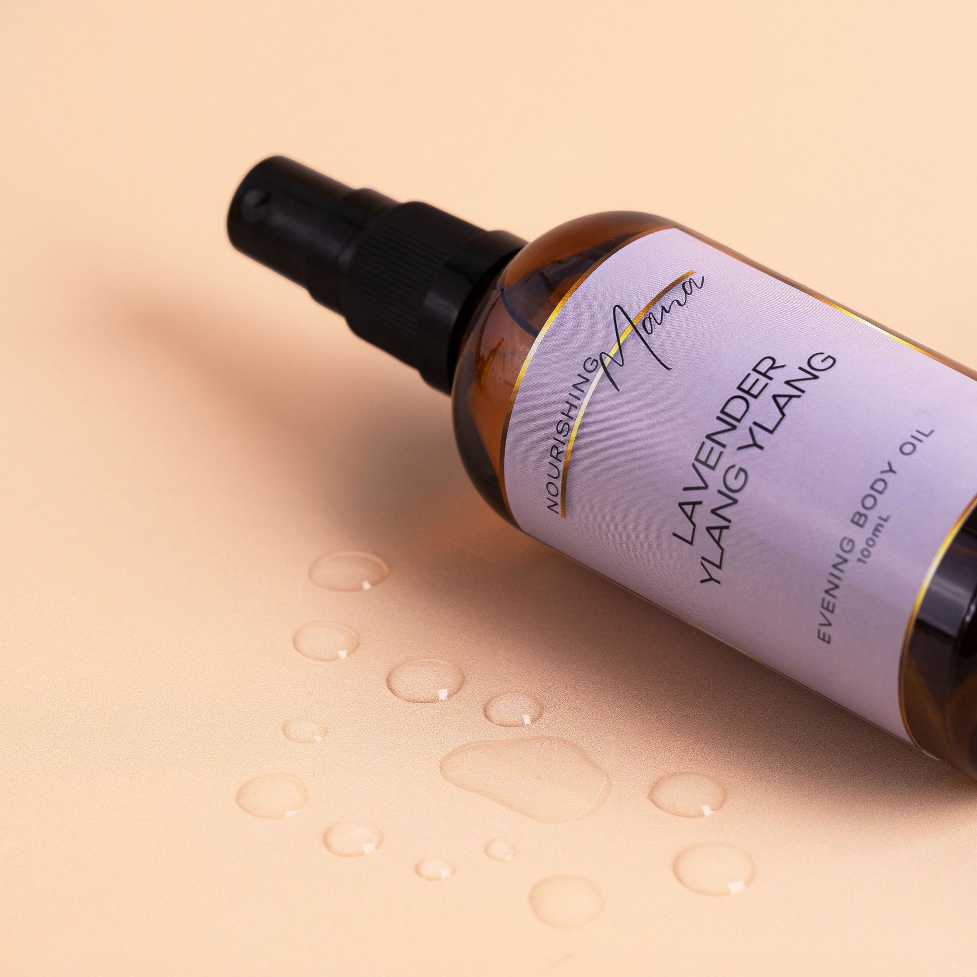 Nourishing Mana Lavender Ylang Ylang Evening Body Oil in a 100ml amber glass spray bottle with oil droplets on a beige background.